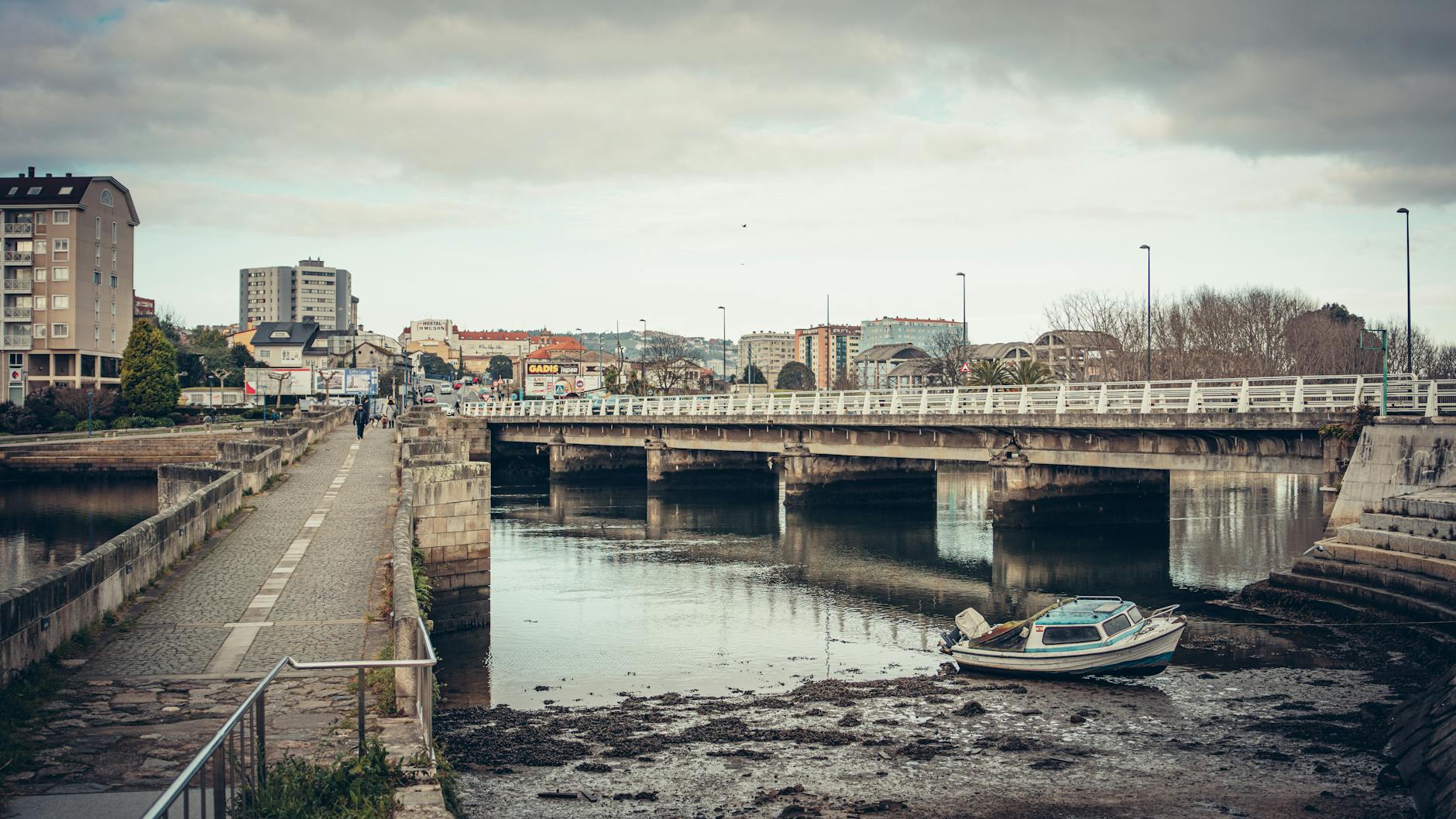 A peaceful urban landscape featuring a bridge over a river in Burgo, Spain, with buildings and a motorboat.