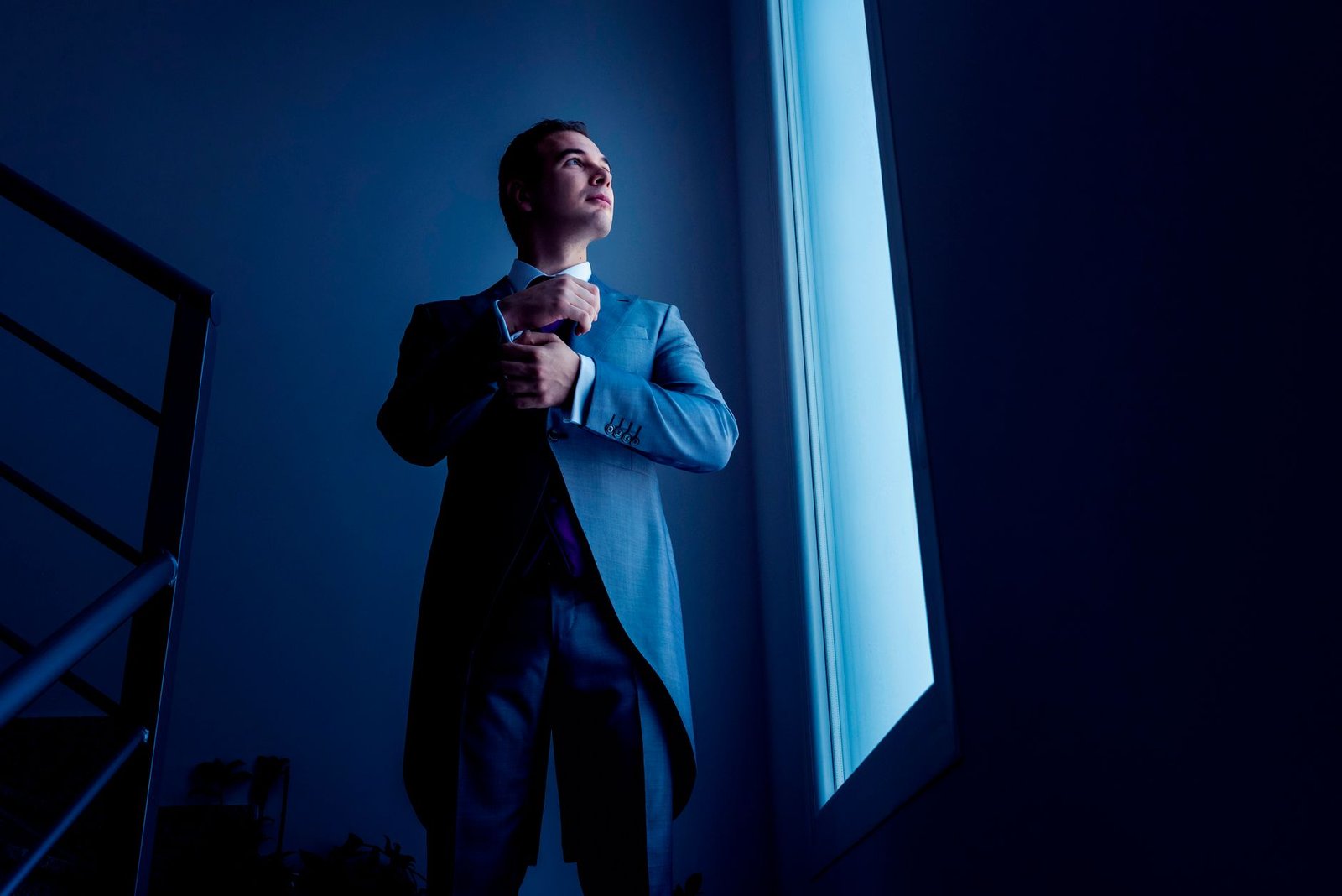 A man in a suit stands near a window with dramatic lighting, evoking mystery and sophistication.